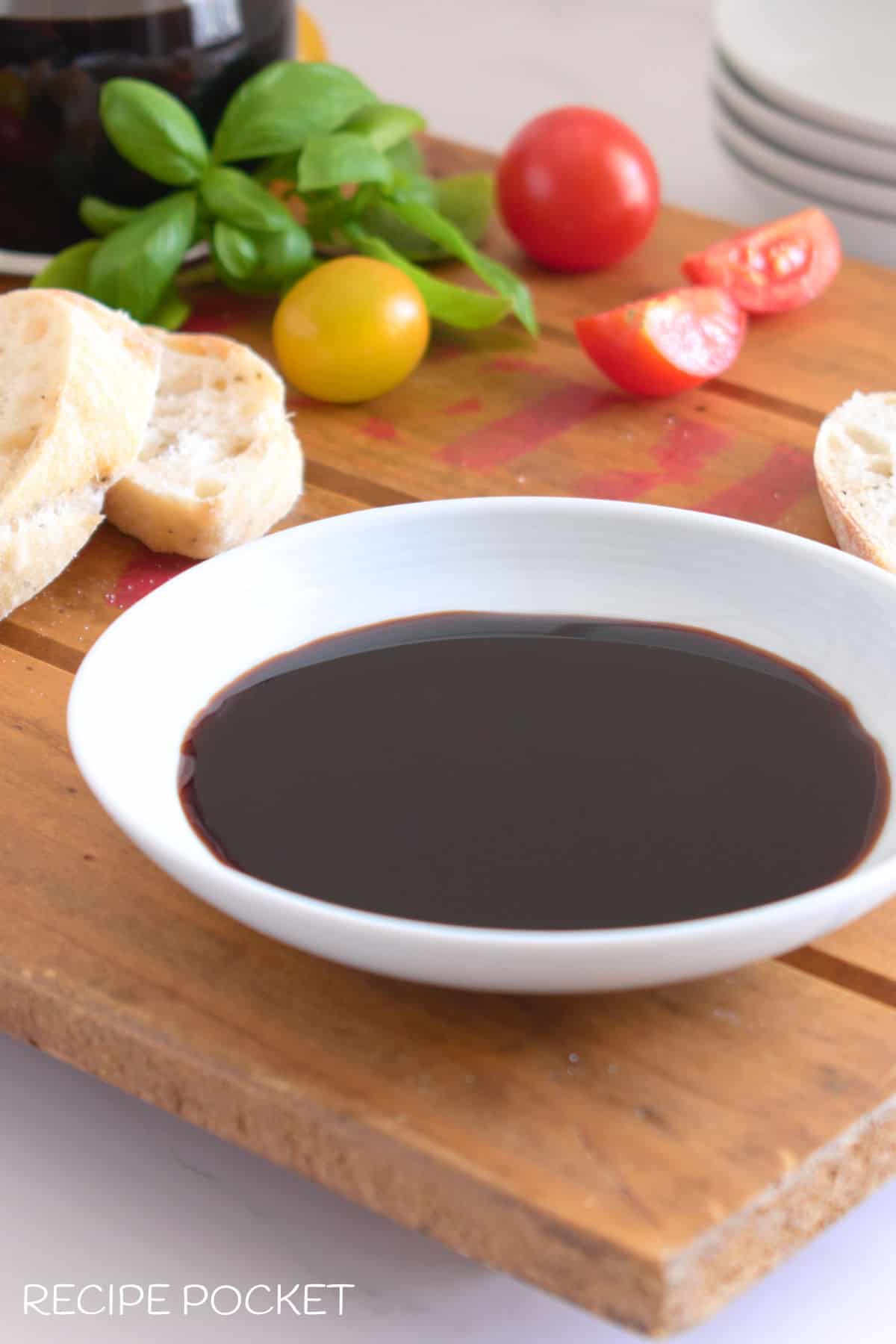 Balsamic vinegar glaze in a white bowl with bread and tomatoes in the background.