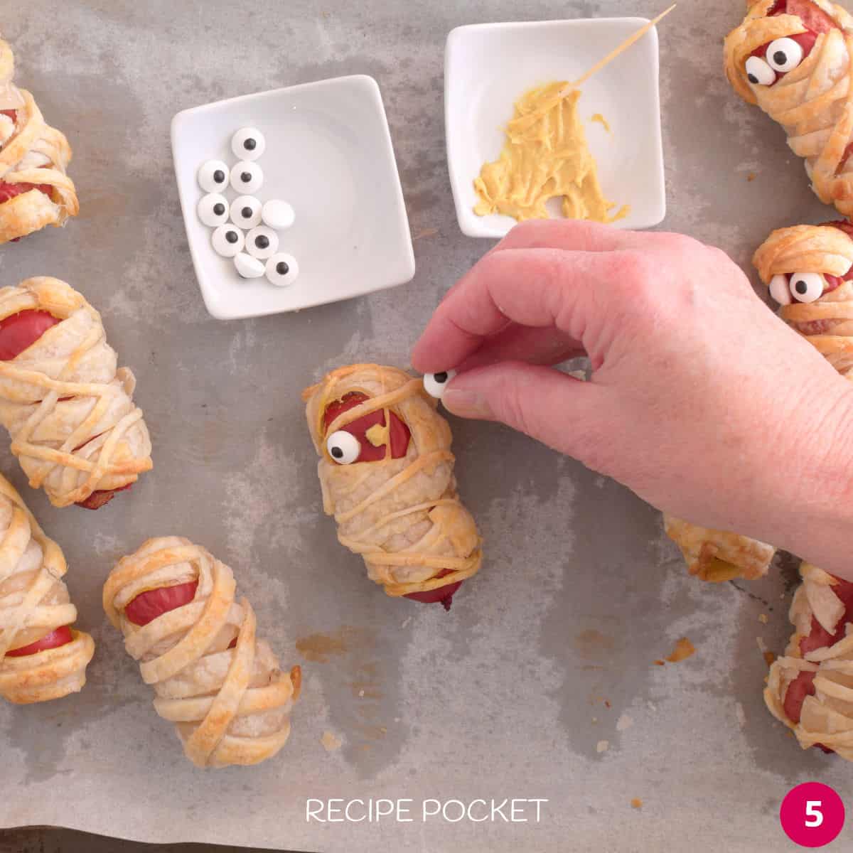 Candy eyes being attached to hot dog mummies.