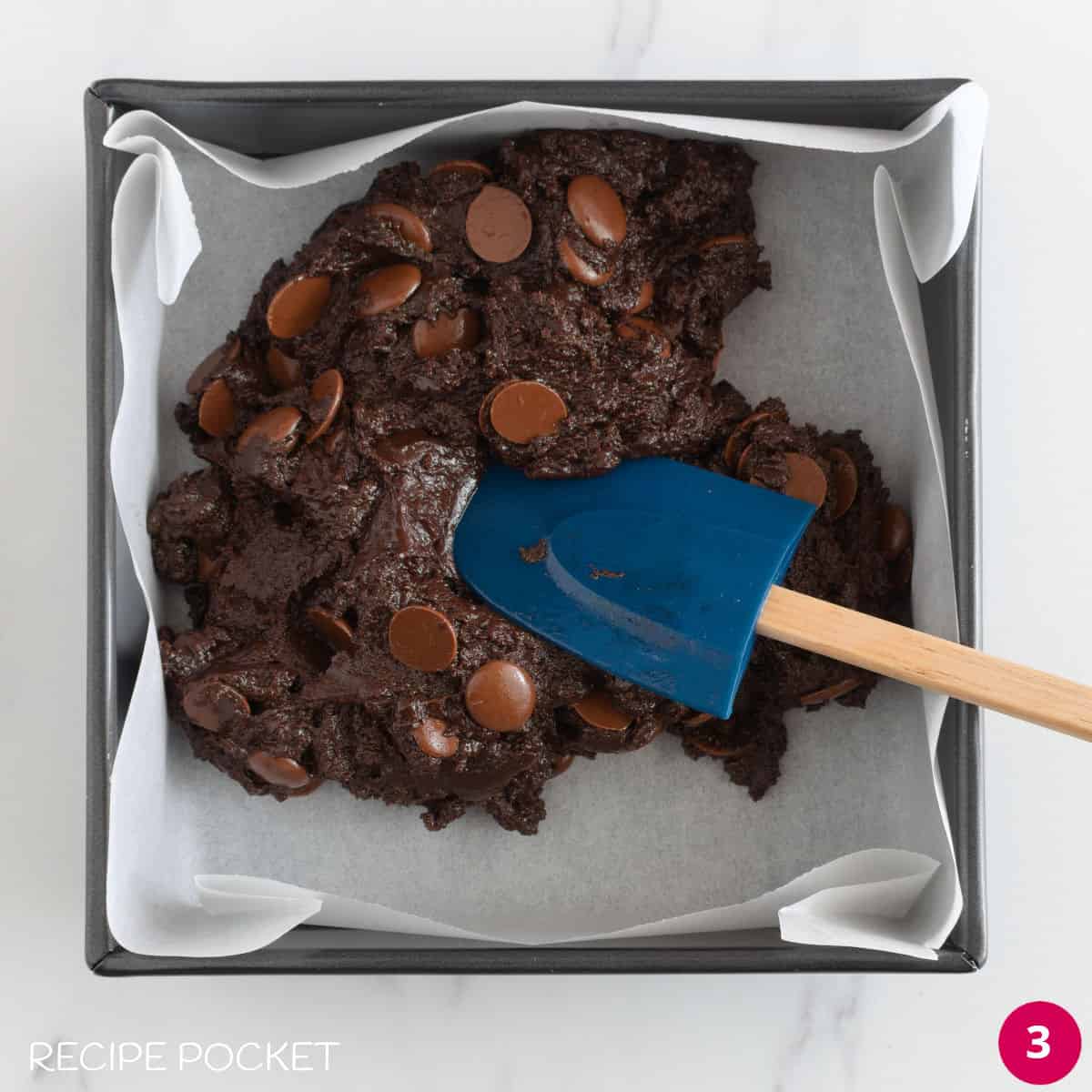 Brownie batter with chocolate chips in a cake tin lined with parchment paper.