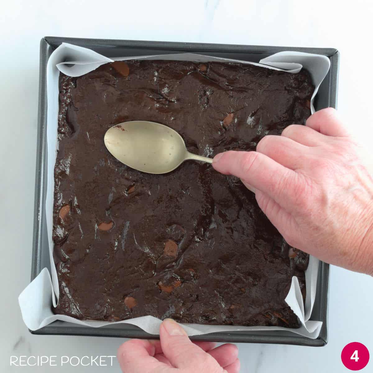 Brownie batter being smoothed evenly in a cake tin with the back of a spoon.