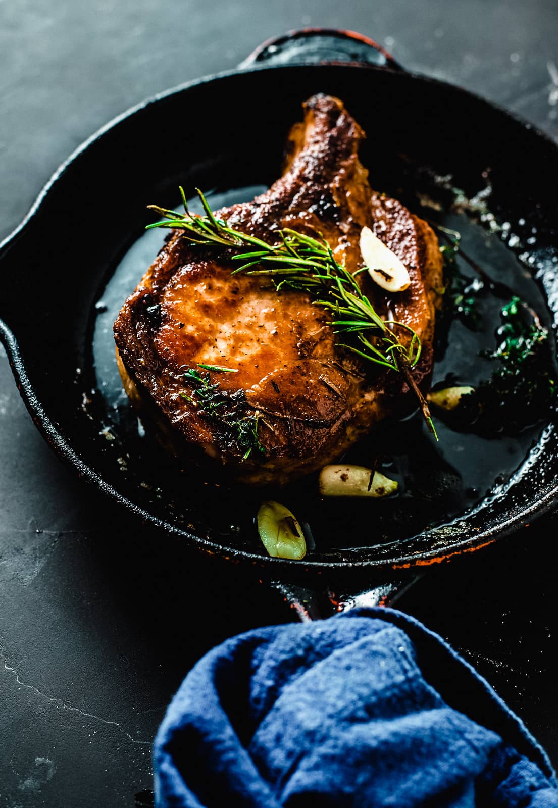 A pork chop with rosemary and garlic in a black skillet.